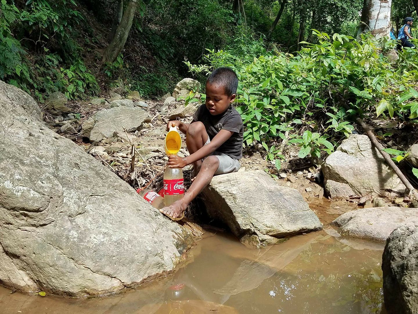 Young boy collecting dirty water from a stream into a Coke bottle
